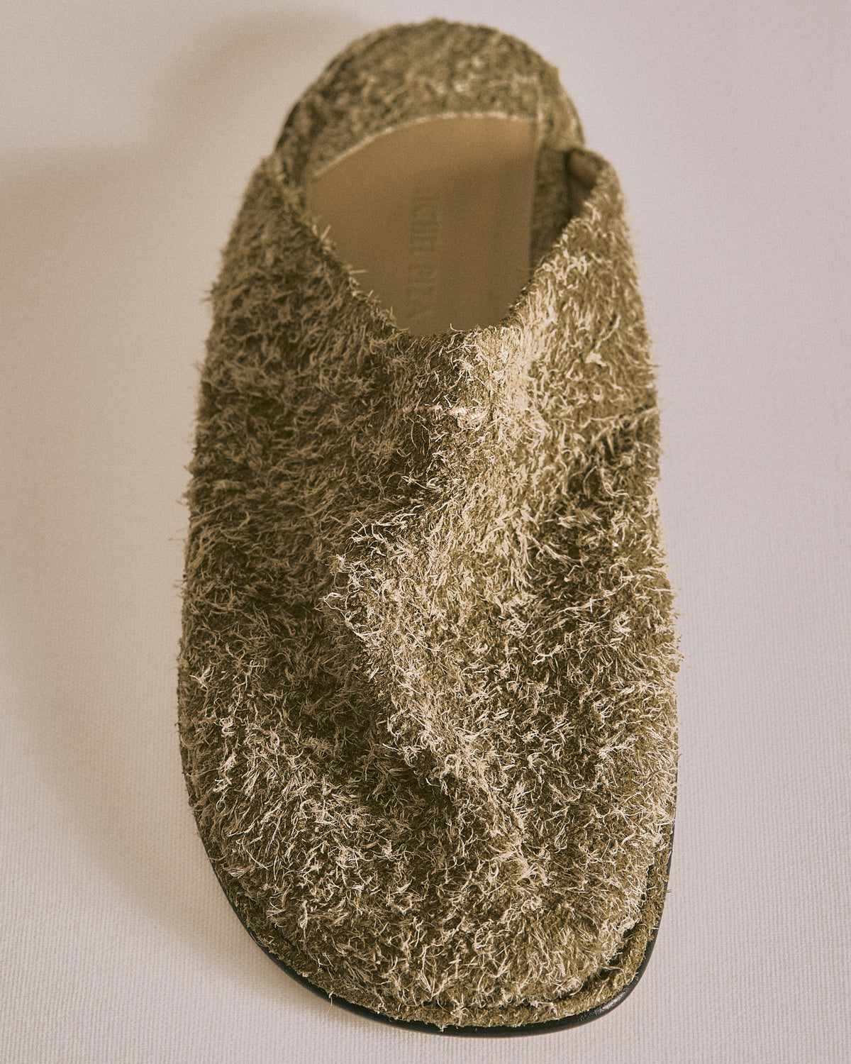 A single fuzzy slip-on mule shoe in a textured olive-brown fabric, photographed from a top-down angle on a light neutral background.